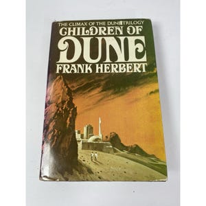 May include: A vintage paperback book titled "Children of Dune" by Frank Herbert. The cover features a desert landscape with a city and two figures, set against an orange and brown background. The title is in large, bold white letters.