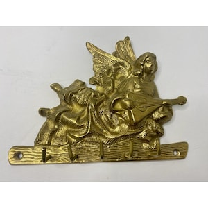 May include: A brass-colored wall-mounted key holder featuring an angel playing a stringed instrument. The angel has detailed wings and is positioned above a horizontal bar with three hooks for keys. The key holder has a vintage aesthetic.