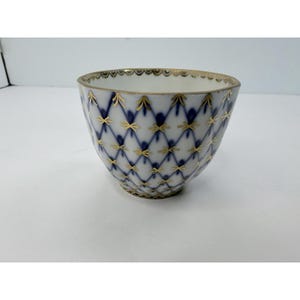 May include: A white porcelain bowl featuring a blue lattice pattern overlaid with gold star-like accents. The rim is decorated with a gold scalloped edge, and the exterior surface displays a repeating diamond grid design in blue and gold.