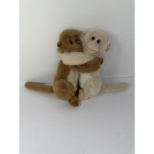 May include: Two plush monkey toys in a hugging pose. One monkey is dark brown with a lighter tan face and chest, while the other is cream-colored with a small pink bow near its ear. Both have dark plastic eyes and long, slender tails.