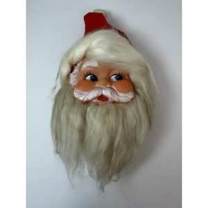 May include: A vintage Santa Claus face decoration with a red hat and a long, white beard. The face is made of plastic with blue eyes, rosy cheeks, and a red mouth. The beard is made of fluffy, white material. A festive holiday decoration.