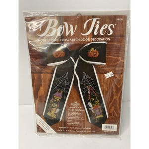 May include: A cross-stitch door decoration kit featuring a black bow tie design with embroidered pumpkins and spiderwebs. The kit includes the text "Bow Ties" and "The Haunted House". Finished size is 11" x 17" (28 cm x 43 cm).