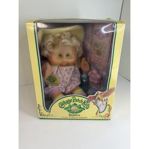 May include: A Cabbage Patch Kids baby doll in its original box. The doll has blonde hair, green eyes, and is wearing a pink and white patterned outfit. The box is yellow with the Cabbage Patch Kids logo and the words "Babies" and "The One of a Kind, For Adoption!"