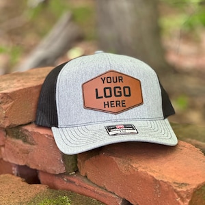 May include: Gray and black trucker hat with a brown leather patch that says "YOUR LOGO HERE".