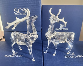 May include: Two crystal reindeer figurines with white antlers. The reindeer are standing on blue boxes with the Swarovski logo.