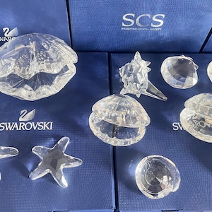Può includere: Una collezione di figurine di cristallo trasparente a forma di animali marini, tra cui una conchiglia, una stella marina, conchiglie e un cavalluccio marino. Le figurine sono disposte su una superficie di velluto blu con il logo Swarovski.