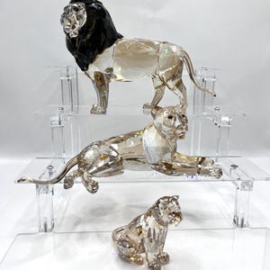 May include: Three crystal lion figurines on clear acrylic shelves. The top lion has a black mane. The middle lion is lying down, and the bottom lion is seated. The figurines are faceted, reflecting light.