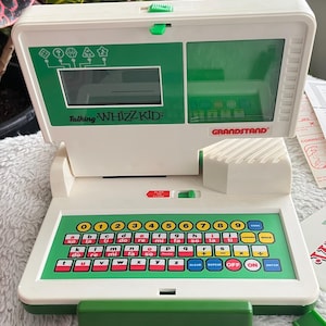 Può includere: Un giocattolo educativo elettronico vintage Talking Whizz Kid. Il giocattolo è bianco e verde, con una tastiera, uno schermo e il logo "GRANDSTAND". La tastiera ha lettere, numeri e note musicali. Il giocattolo è progettato per scopi educativi.