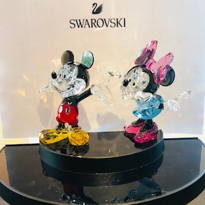 May include: Crystal figurines of Mickey and Minnie Mouse. Mickey wears red shorts, yellow shoes, and has black ears. Minnie wears a blue skirt, pink shoes, and a pink bow. Both figures are on a black display stand, with the Swarovski logo in the background.