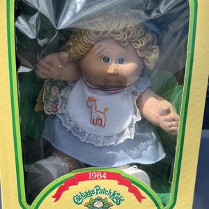 Puede incluir: Muñeca vintage Cabbage Patch Kids en su caja original. La muñeca tiene cabello rubio rizado, ojos azules y un babero blanco con un diseño de jirafa. La caja es amarilla y verde con el texto "Cabbage Patch Kids" y "Ptits Bouts de Choux".