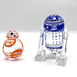 May include: Two crystal figurines of Star Wars characters BB-8 and R2-D2. BB-8 is orange and white, while R2-D2 is blue and white. Both figures are made of faceted crystal, reflecting light.
