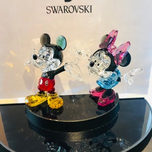 May include: Crystal figurines of Mickey and Minnie Mouse. Mickey wears red shorts, yellow shoes, and has a black body. Minnie has a pink bow, blue dress, and pink shoes. The figurines are on a black display stand, with the Swarovski logo in the background.