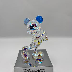 May include: A crystal figurine of Mickey Mouse in a standing pose with blue ears and a black nose. The figurine is on a silver base with the text "Disney 100" printed on it.