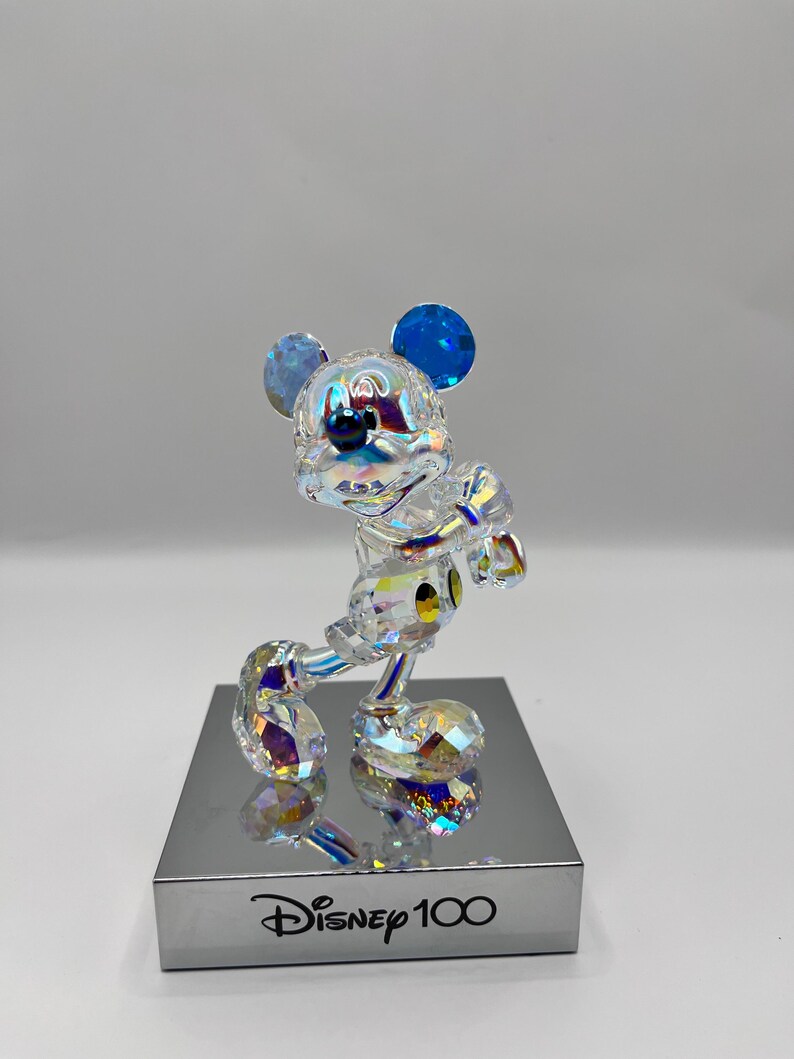 May include: A crystal figurine of Mickey Mouse in a standing pose with blue ears and a black nose. The figurine is on a silver base with the text "Disney 100" printed on it.