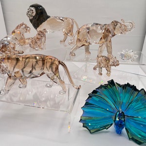 Op de afbeelding: Een verzameling kristallen dierenfiguren, waaronder een leeuw, een tijger, een olifant en een pauw. De figuren zijn allemaal in tinten bruin, blauw en groen.