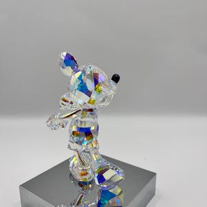 May include: A crystal figurine of Mickey Mouse standing on a silver base. The figurine is iridescent and has a black nose.