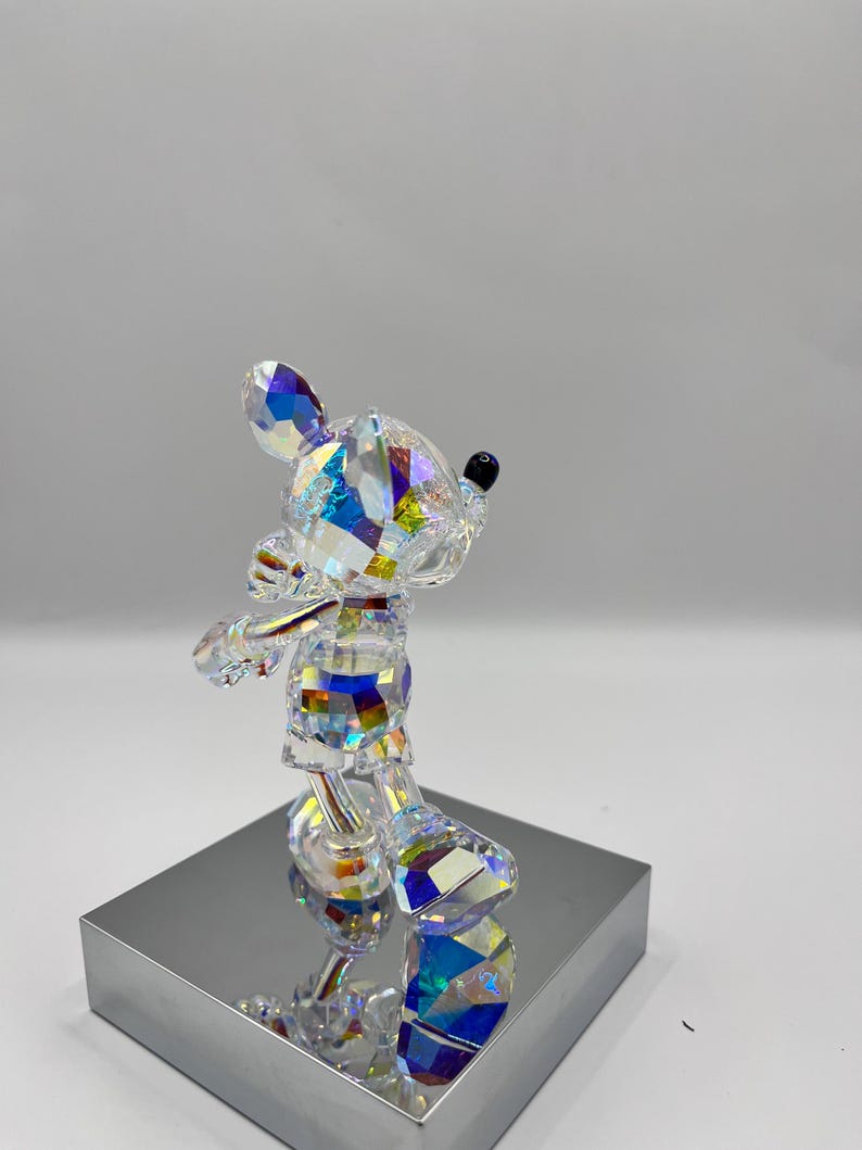 May include: A crystal figurine of Mickey Mouse standing on a silver base. The figurine is iridescent and has a black nose.
