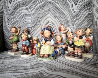 Op de afbeelding: Een verzameling Hummel-beeldjes die kinderen in verschillende poses uitbeelden. De beeldjes hebben gedetailleerde kleding en accessoires, met een focus op traditionele Duitse ontwerpen. Het kleurenpalet omvat pasteltinten en aardetinten, waarbij elk beeldje op een kleine basis is gemonteerd.