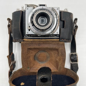 May include: A silver vintage camera with a black leather case and strap. The camera has a lens with a silver ring that reads "Solinar 1:3.5 42.50 AGFA" and "COMPUR-RAPID". The camera is in good condition and has a classic look.