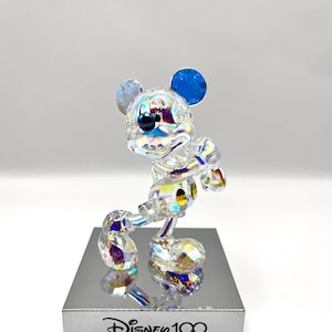 May include: A crystal figurine of Mickey Mouse in a standing pose with his signature yellow gloves and blue ears. The figurine is iridescent and has a reflective base with the text "Disney 100" printed on it.