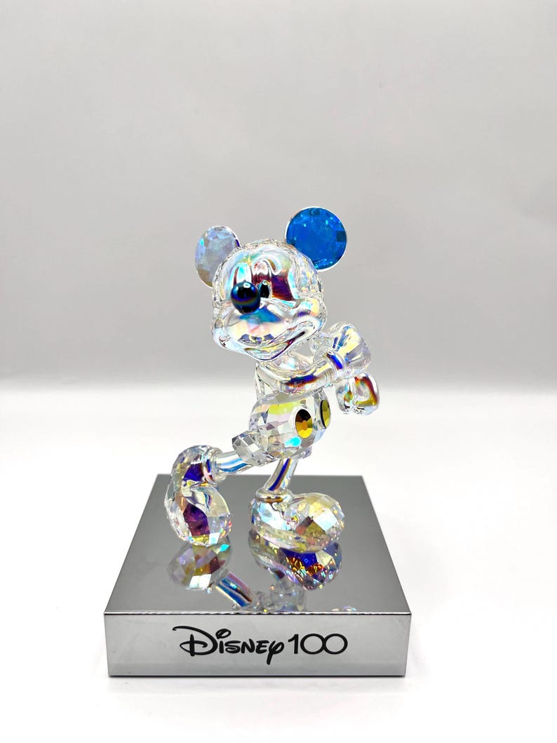 May include: A crystal figurine of Mickey Mouse in a standing pose with his signature yellow gloves and blue ears. The figurine is iridescent and has a reflective base with the text "Disney 100" printed on it.