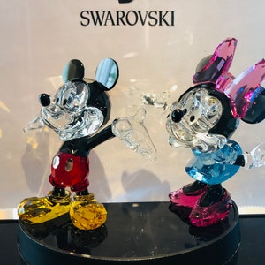 May include: Crystal figurines of Mickey and Minnie Mouse. Mickey is wearing red shorts, yellow shoes, and black gloves. Minnie is wearing a blue dress, pink shoes, and a pink bow. The figurines are on a black base, with the Swarovski logo in the background.