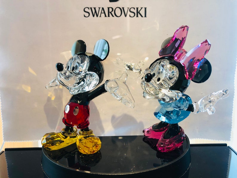 May include: Crystal figurines of Mickey and Minnie Mouse. Mickey is wearing red shorts, yellow shoes, and black gloves. Minnie is wearing a blue dress, pink shoes, and a pink bow. The figurines are on a black base, with the Swarovski logo in the background.