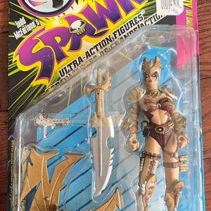 May include: A Spawn Ultra-Action Figure, packaged in a clear plastic blister pack. The figure is of a female warrior with gold armor and a large sword. The packaging includes the text "Todd McFarlane's Spawn".