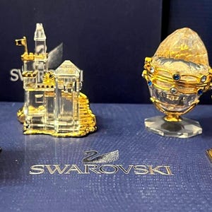 May include: Four crystal figurines displayed on a blue surface. The figurines are a castle, a globe, a temple, and a pagoda. Each figurine is made of clear crystal with gold accents. The figurines are all approximately 7.5 cm tall. The surface is blue and has the words "Swarovski" and "SCS" printed on it.