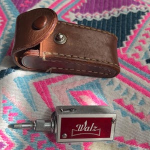 May include: A rectangular silver-toned metal device with a red panel featuring the white text Walz, accompanied by a brown leather carrying case with a snap button closure. The device includes a small metal adjustment knob on one side.