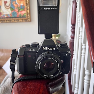 Puede incluir: Cámara de película Nikon N2000 negra con flash Nikon SB-E. La cámara tiene una lente de 50 mm 1:1.8. La correa de la cámara es negra. Está sobre una superficie de madera.