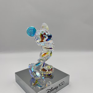 May include: A crystal figurine of Mickey Mouse in a standing pose. The figurine is clear with iridescent accents and blue ears. The figurine is on a silver base with the text "Disney 100" printed on it.