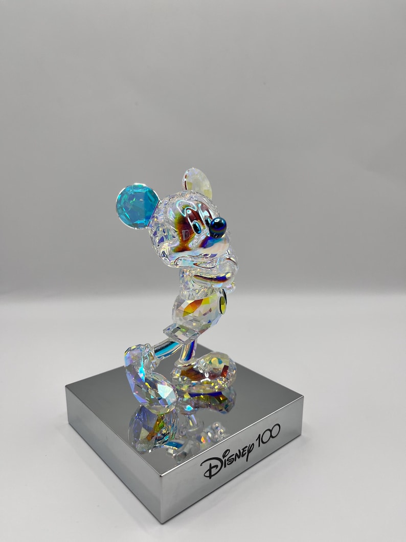 May include: A crystal figurine of Mickey Mouse in a standing pose. The figurine is clear with iridescent accents and blue ears. The figurine is on a silver base with the text "Disney 100" printed on it.