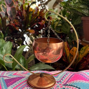 May include: A polished copper bowl suspended by three thin metal chains from a curved brass-coloured stand. The stand features a circular base and a hook at the top to hold the bowl. The copper bowl has a smooth, reflective, rounded exterior.