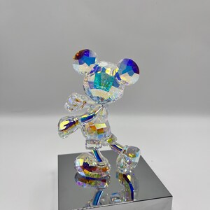 May include: A crystal figurine of Mickey Mouse standing on a silver base. The figurine is iridescent and has a rainbow effect.