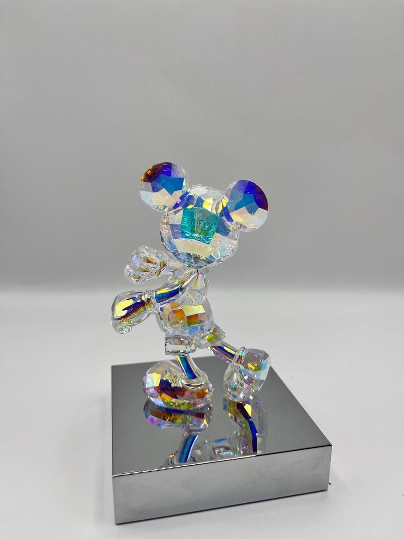 May include: A crystal figurine of Mickey Mouse standing on a silver base. The figurine is iridescent and has a rainbow effect.