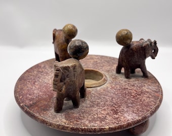 May include: A handcrafted, circular, reddish-brown stone sculpture featuring three small elephant figures. Each elephant has a small, round ball balanced on its back. The sculpture has a central recessed area and is set on a low base.