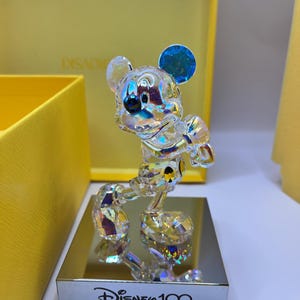 May include: A crystal figurine of Mickey Mouse in a standing pose, with a blue iridescent ear. The figurine is on a silver base with the text "Disney 100" inscribed on it.