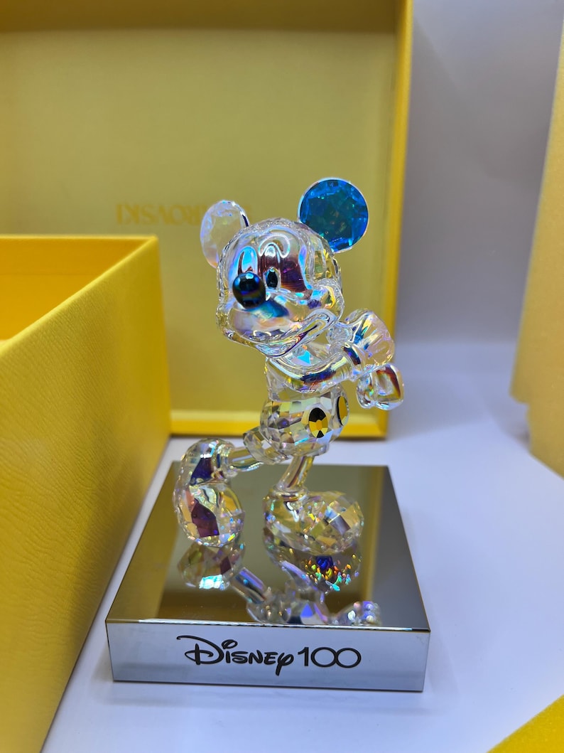 May include: A crystal figurine of Mickey Mouse in a standing pose, with a blue iridescent ear. The figurine is on a silver base with the text "Disney 100" inscribed on it.