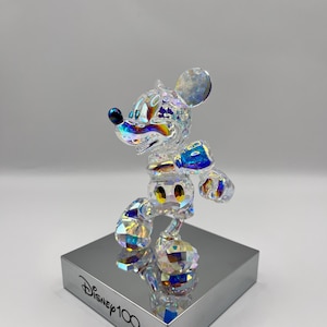 May include: A crystal figurine of Mickey Mouse in a standing pose. The figurine is iridescent and features a blue bow tie and yellow buttons. The figurine is on a silver base with the text "Disney100" printed on it.