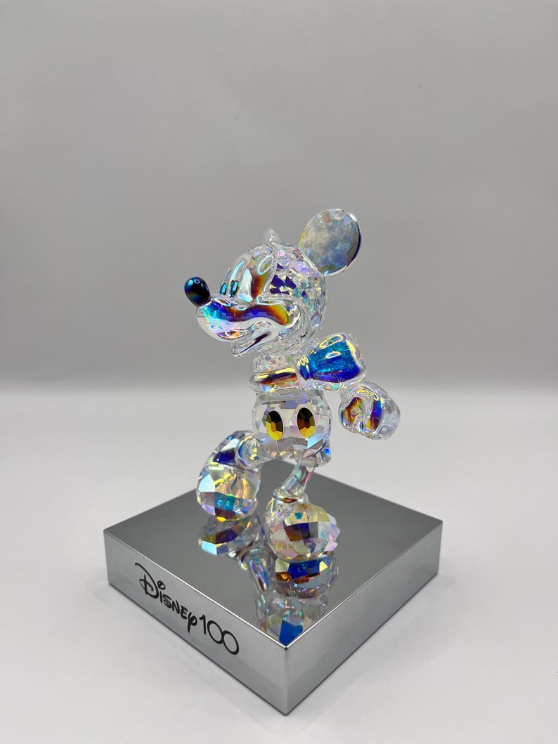 May include: A crystal figurine of Mickey Mouse in a standing pose. The figurine is iridescent and features a blue bow tie and yellow buttons. The figurine is on a silver base with the text "Disney100" printed on it.