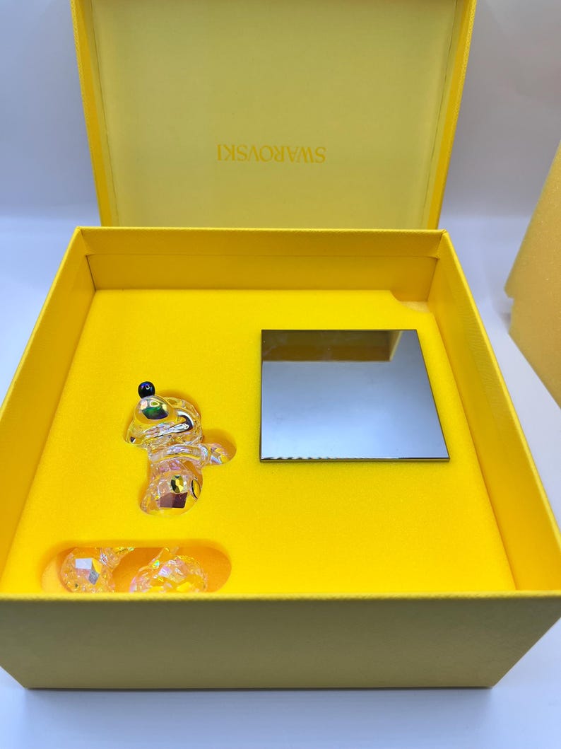 May include: A yellow box with a crystal figurine of a bear inside. The box is open and the bear is sitting on a yellow base. The box has the word "SWAROVSKI" printed on the inside of the lid.