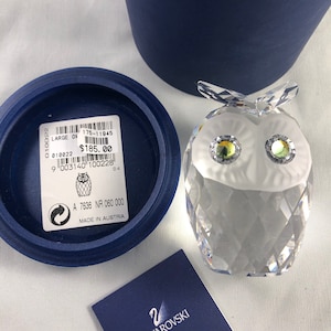 Peut inclure: Une figurine de hibou en cristal transparent avec des détails facettés, des yeux irisés et une poitrine givrée blanche. Le hibou est à côté d'une boîte bleue et d'un certificat Swarovski. Une étiquette avec le texte "MADE IN AUSTRIA" est également visible.