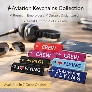 Op de afbeelding: Collectie luchtvaart sleutelhangers met geborduurde labels in diverse kleuren. Sleutelhangers met de woorden "CREW", "PILOT", "I LOVE FLYING" en "I'D RATHER BE FLYING". Zonnebril en koptelefoon op de achtergrond.