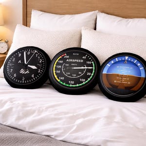 May include: Three circular cushions with aviation-themed designs. One is a clock, another an airspeed indicator, and the third a flight instrument. They are black with white, yellow, green, and blue accents, on a white bed.