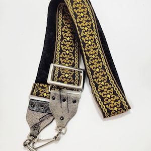May include: A black and gold patterned bag strap with silver-toned hardware. The strap features a velvet backing and decorative gold detailing. The strap has silver metal clasps and a buckle for adjustment.
