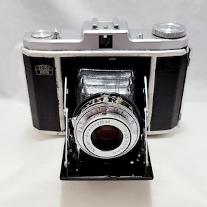 May include: A silver and black Zeiss Ikon camera with a Novar Anastigmat lens. The lens has a maximum aperture of f/4.5 and a focal length of 75mm. The camera has a rangefinder and a built-in flash.