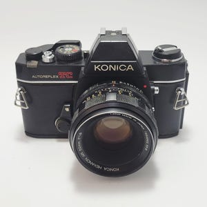 May include: A black Konica Autoreflex TC 35mm film camera with a 52mm Hexanon lens. The camera has a silver top and a black leather body. The lens has a maximum aperture of f/1.8.