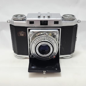 May include: A vintage Zeiss Ikon camera with a black leather body and silver accents. The lens is labeled "Novar-Anastigmat" and "Pronto". The camera has a retractable lens cover and two dials on top.