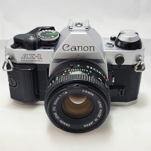 May include: A silver and black Canon AE-1 Program camera with a 50mm lens. The camera has the Canon logo and AE-1 Program text on the body. The lens has green and white markings. The camera is on a white surface.